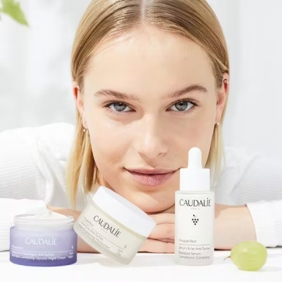 Produtos Caudalie de cuidados de pele em frascos branco e lilás com texto, sobre mesa branca com uva verde e mulher loira ao fundo
