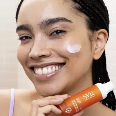 Mulher aplicando creme no rosto segurando frasco laranja SVR