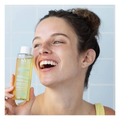 Mulher sorridente a segurar um frasco de líquido amarelo com etiqueta branca em fundo de azulejos azuis