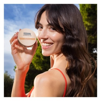 Mulher segurando creme facial ISDIN Eryfotona Actinica em embalagem bege com texto em preto e laranja, ao ar livre com céu azul e árvores.