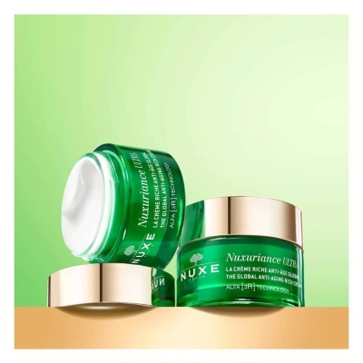 Frascos verdes translúcidos de creme NUXE Nuxuriance Ultra com tampas douradas em superfície bege