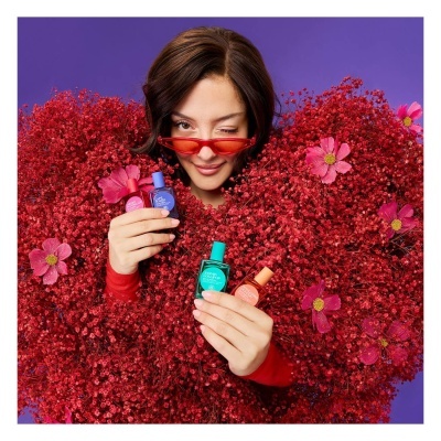 Mulher com óculos vermelhos segurando frascos coloridos de perfumes entre flores vermelhas