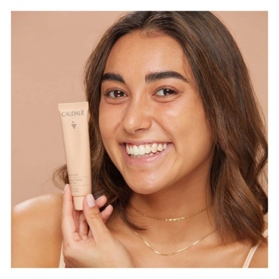 Mulher a segurar tubo do produto CAUDALIE, fundo bege claro.