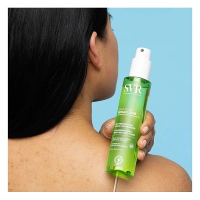 Frasco verde translúcido de spray corporal SVR SEBIACLEAR sobre o ombro de uma mulher com fundo azul.