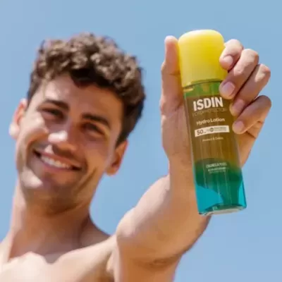 Frasco ISDIN Hydro Lotion com tampa amarela segurado por homem sob céu azul