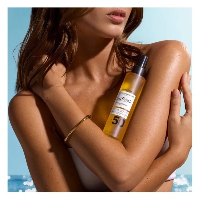 Frasco de protetor solar Lierac SPF 50 segurado por mulher bronzeada com fundo de mar azul