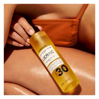 Frasco amarelo de óleo solar Lierac FPS 30 segurado sobre perna bronzeada