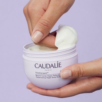 Creme corporal Caudalie em frasco branco com creme branco sendo retirado pelos dedos