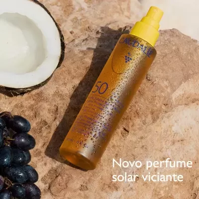 Frasco amarelo de spray solar FPS 50 da CAUDALIE com gotas de água sobre rocha junto a coco e uvas.
