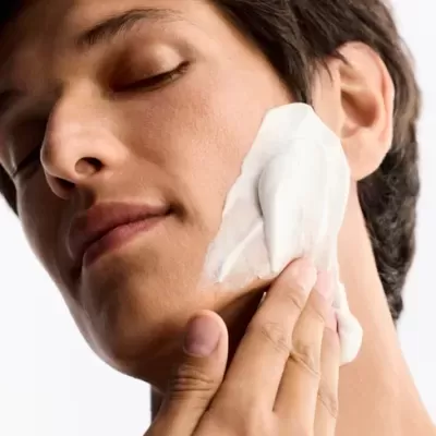 Pessoa aplicando creme facial branco no rosto com olhos fechados