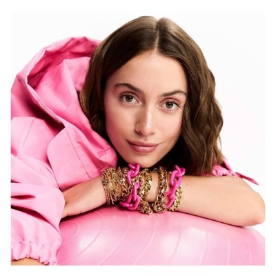 Mulher com blusa rosa e pulseiras douradas e rosa apoiada num objeto rosa