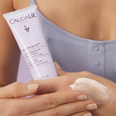 Creme para mãos e unhas Caudalie sendo aplicado na mão de uma pessoa com vestido azul claro
