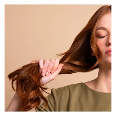 Mulher com cabelo ruivo comprido e ondulado a segurar cabelo, veste camisola verde-militar