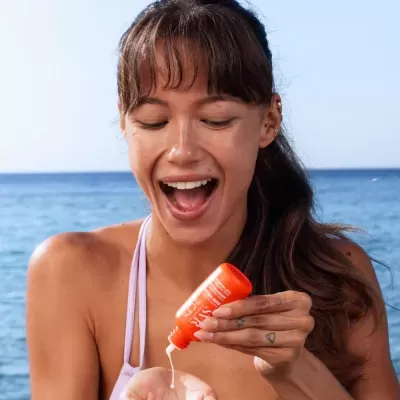 Mulher a colocar protetor solar na mão com frasco laranja à beira-mar