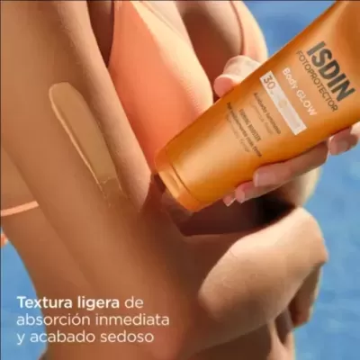 Mulher com fato de banho laranja aplica protetor solar ISDIN Body Glow SPF 50+ no braço com fundo azul.