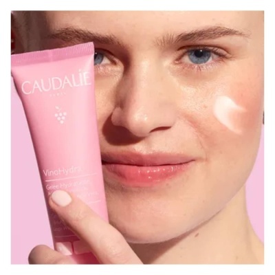 Mulher segurando creme facial rosa CAUDALÍE VinoHydra com creme aplicado na bochecha