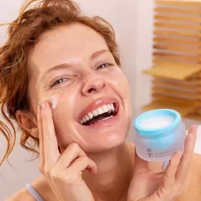 Mulher aplicando creme facial de pote azul e branco com janela de persianas ao fundo
