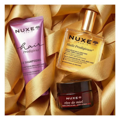 Produtos cosméticos Nuxe: champô rosa, óleo multiusos em frasco e bálsamo labial em pote, sobre tecido dourado