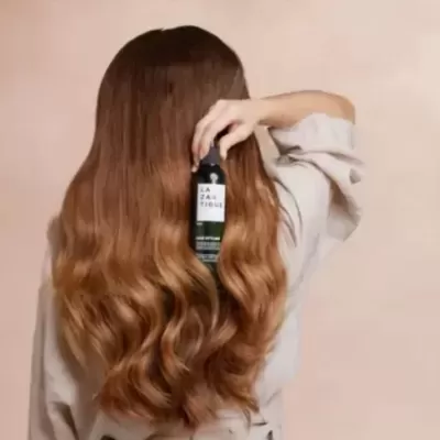 Mulher com cabelo castanho longo e encaracolado segurando garrafa preta de produto LAZARIDIQUE
