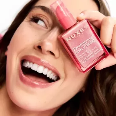Frasco rosa metálico do sérum NUXE Prodigieuse Hyalu Boost perto do rosto de uma mulher sorridente