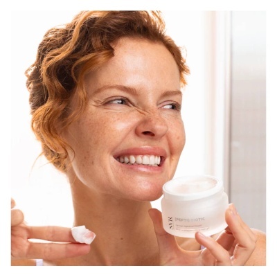 Mulher segurando frasco de creme facial branco fosco enquanto aplica produto com dedo num ambiente iluminado naturalmente