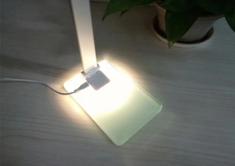 CANDEEIRO LED - DESIGN MODERNO Lâmpada de mesa branca com base transparente sobre mesa de madeira clara junto a planta verde