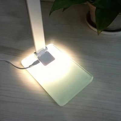 Lâmpada de mesa branca com base transparente sobre mesa de madeira clara junto a planta verde
