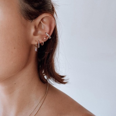 Lado esquerdo do rosto com vários brincos pequenos de metal e colar dourado com pendente