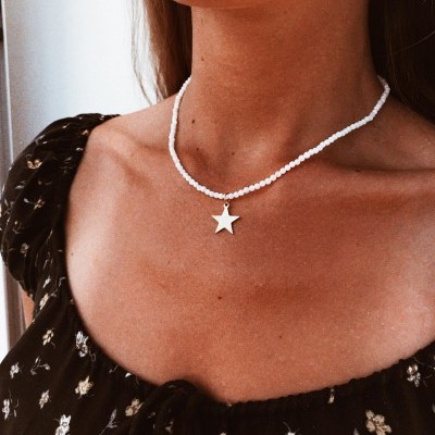 Colar com pendente de estrela prateada e contas brancas num pescoço feminino, blusa preta com padrão floral.