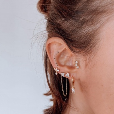 Orelha feminina com brincos dourados e pedras brancas em fundo branco.