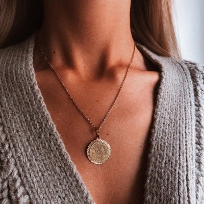 Colar dourado com pendente redondo espiral sobre suéter cinza de malha