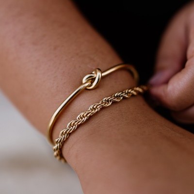 Pulso com duas pulseiras douradas de metal, uma com nó e outra de corrente entrelaçada