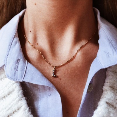 Colar dourado com letra L em pedras pretas no pescoço de pessoa com camisa azul e casaco branco