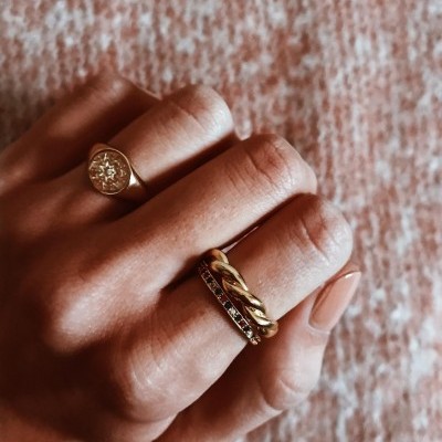 Mão com três anéis dourados distintos, fundo tecido rosa claro.