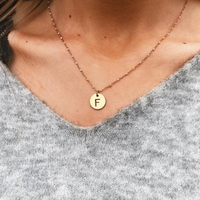 Colar dourado com pendente redondo e letra 'F' usado com camisola cinzenta