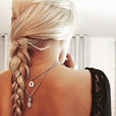 Mulher com cabelo loiro em trança e dois colares prateados com pendentes, de costas.