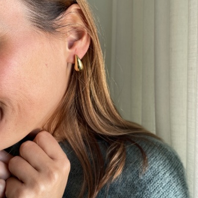 Brinco dourado em forma de gota na orelha de mulher com cabelo loiro e suéter cinza
