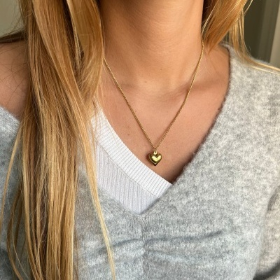 Colar dourado com pendente de coração usado por pessoa com camisola cinza.