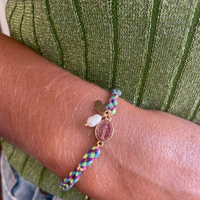 Pulseira trançada colorida com medalhão religioso, pendente de folha e pedra branca numa mão sobre fundo de roupa verde