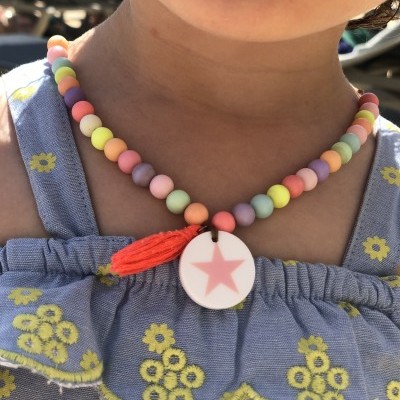 Colar colorido com estrela rosa pendurada em pescoço de criança com vestido azul e flores amarelas.