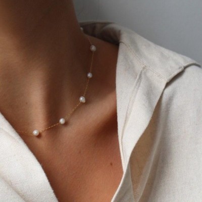 Colar fino dourado com pérolas usado no pescoço e camisa bege clara