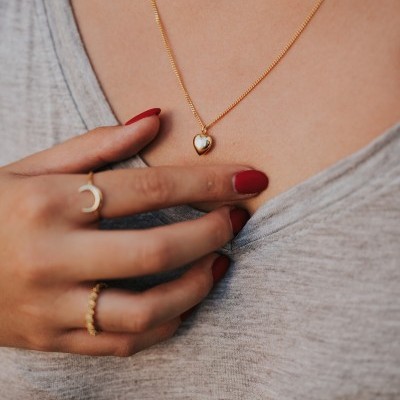 Colar dourado com pendente em forma de coração e duas mãos com anéis dourados e unhas vermelhas sobre camisola cinzenta