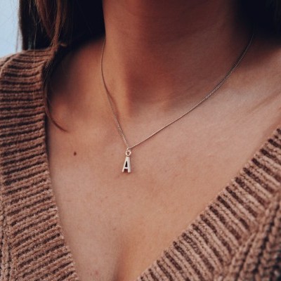 Colar prateado com pendente em forma da letra A no pescoço de uma pessoa com casaco castanho