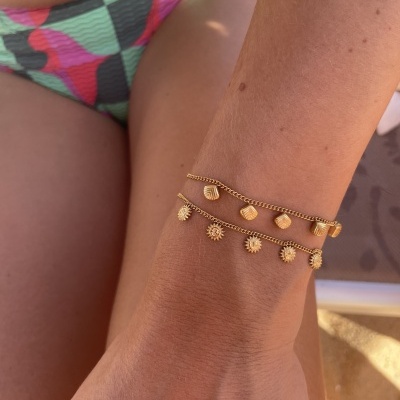 Pulseiras douradas com pingentes de conchas e sóis no pulso de uma pessoa com fato de banho colorido.