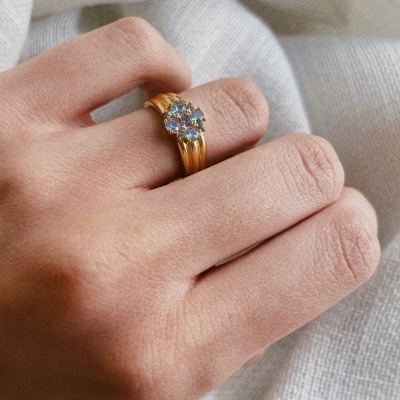 Anel dourado com pedras azuis em formato de flor, na mão sobre tecido branco