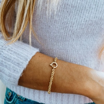 Pulseira dourada com fecho circular no pulso de mulher com camisola cinza e calças de ganga.