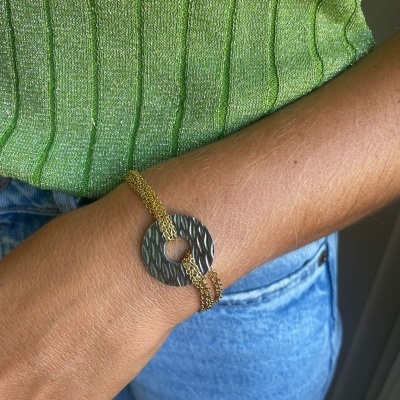 Pulseira com pendente circular texturizado e corrente dourada no pulso de uma pessoa com camisa verde e calças de ganga