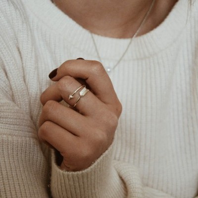 Anel dourado minimalista em dedo de mão com unhas castanho escuro sobre suéter branco