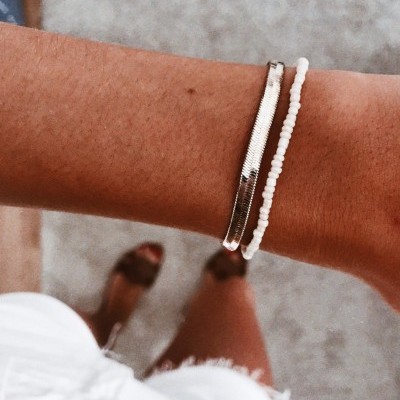 Pulseiras prata e brancas no pulso com fundo de chão cinzento e peça de roupa branca.