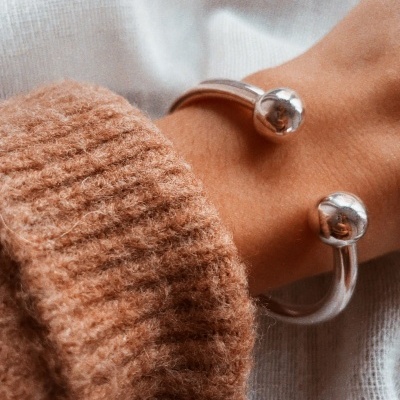 Pulseira prateada aberta com esferas nas extremidades no pulso de uma pessoa com camisola castanha